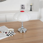 Clérachic table lamp, unlit, on a wooden table beside a checked mug resting on an open magazine, a framed print on the shelf behind.