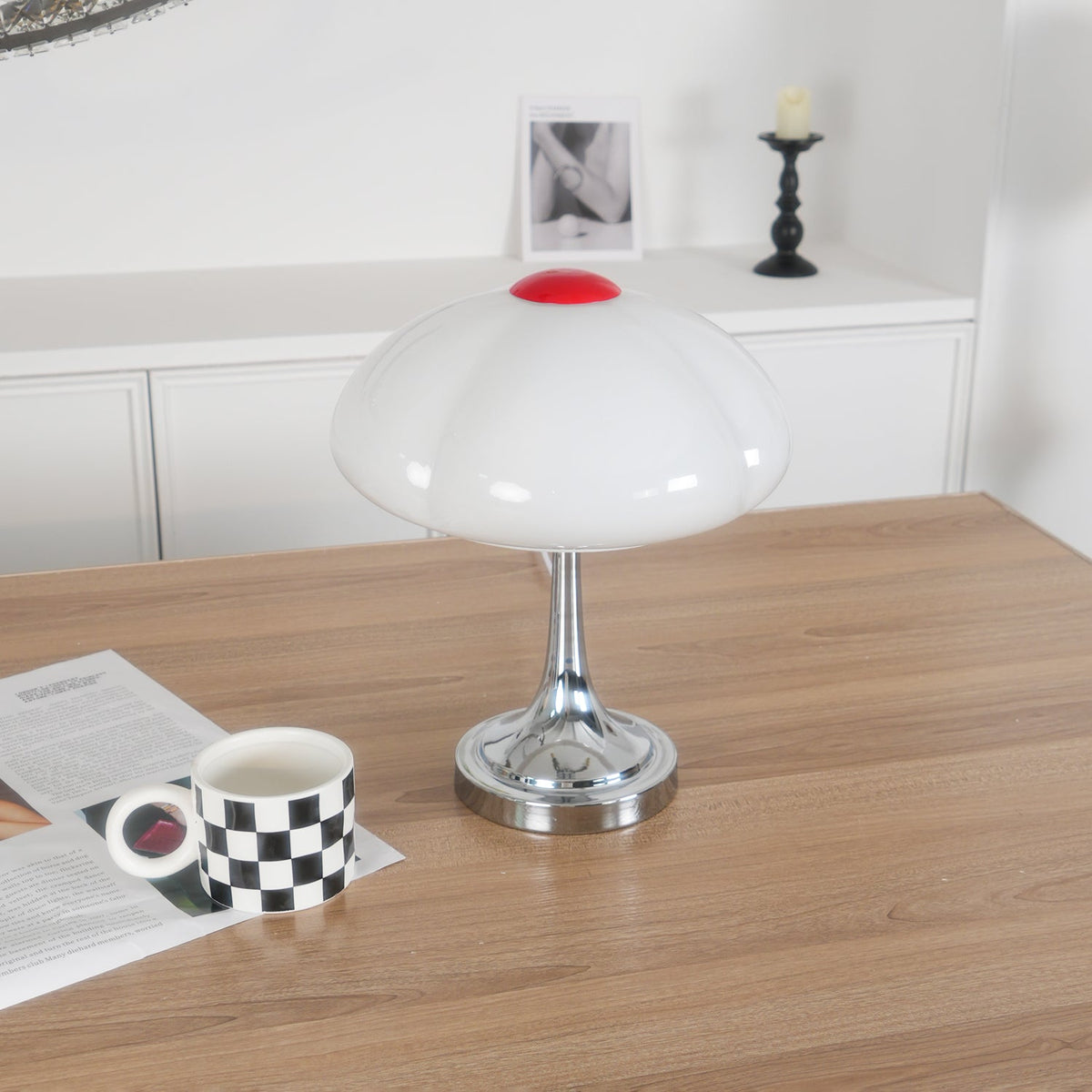 Clérachic table lamp, unlit, on a wooden table beside a checked mug resting on an open magazine, a framed print on the shelf behind.