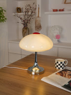 A glowing Clérachic table lamp on a wooden desk, with a checked mug, a magazine and shelves of decor in the background.