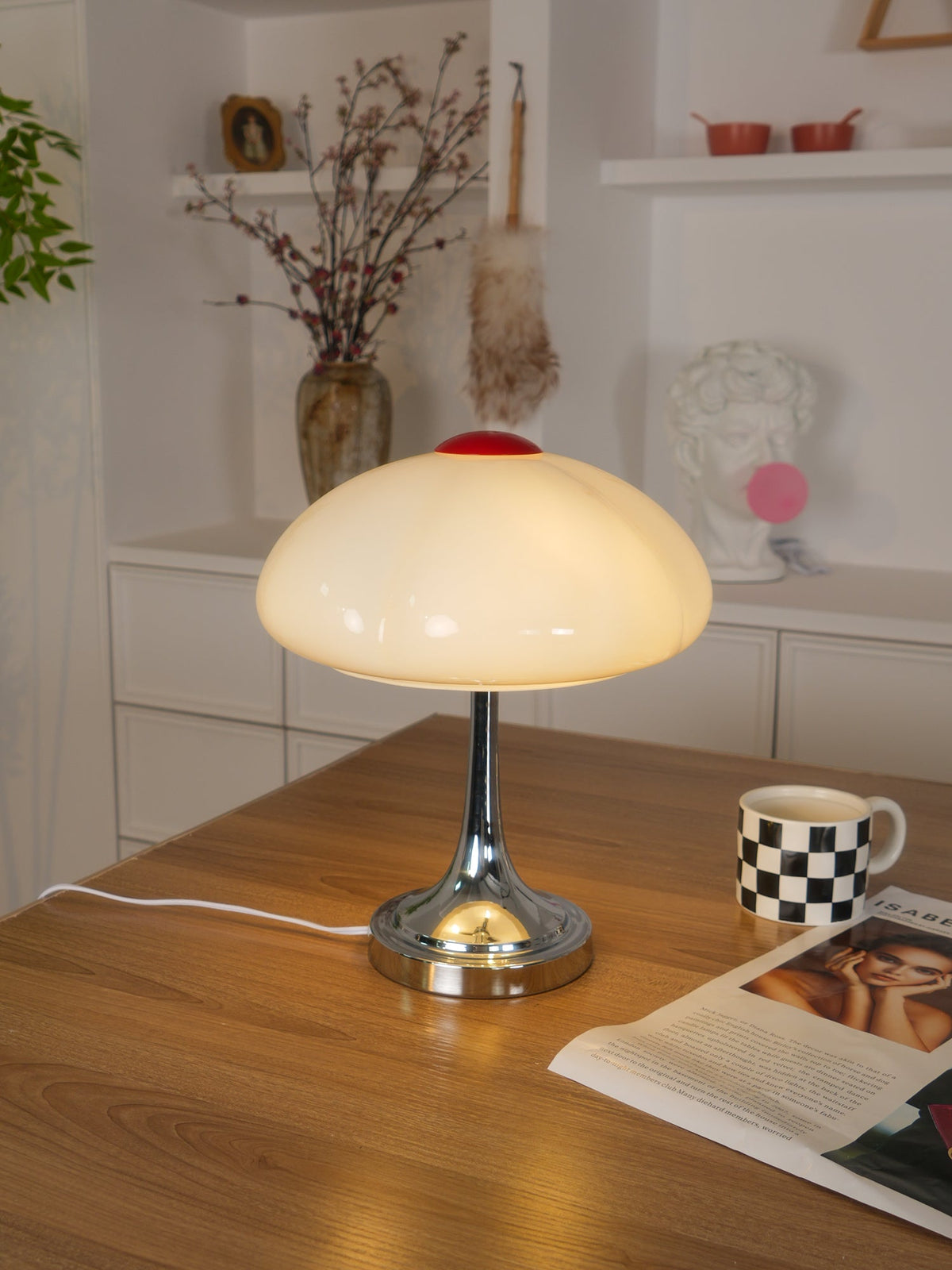 A glowing Clérachic table lamp on a wooden desk, with a checked mug, a magazine and shelves of decor in the background.