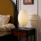 Oval-shaped Clérachic table lamp lit on a dark bedside table, beside a rumpled bed with a mustard-yellow headboard.