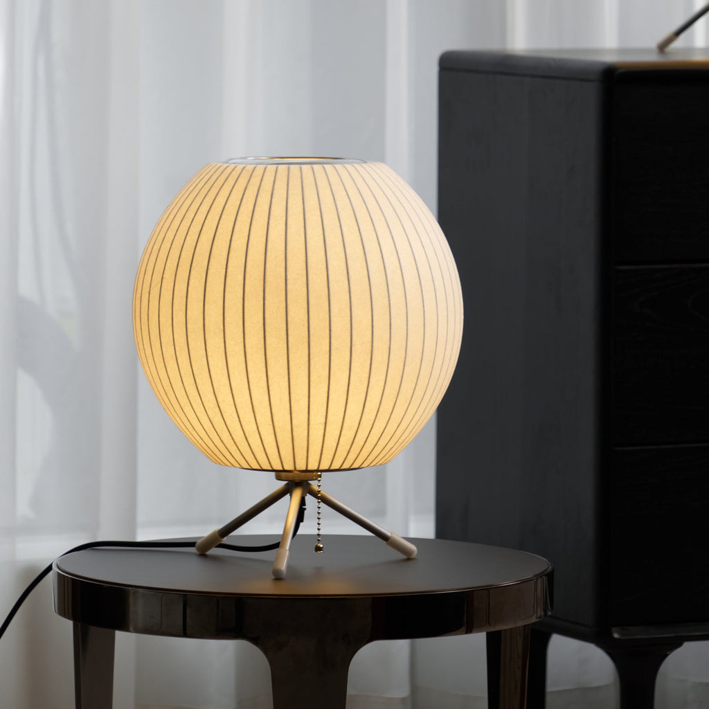 A rounded, glowing Clérachic table lamp on a dark glass-topped side table, with a tall black cabinet and white curtains behind.