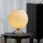 A rounded, glowing Clérachic table lamp on a dark glass-topped side table, with a tall black cabinet and white curtains behind.
