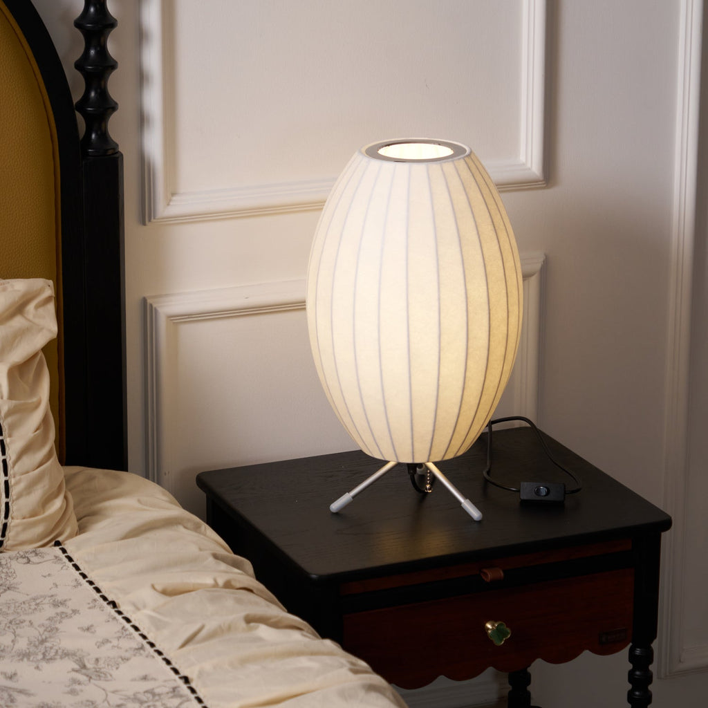 Unlit oval white shade on a small tripod, this Clérachic table lamp stands on a dark bedside table beside a cream bed.
