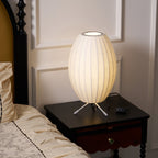 Unlit oval white shade on a small tripod, this Clérachic table lamp stands on a dark bedside table beside a cream bed.