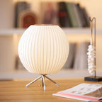 Soft focus bookshelves frame a round white Clérachic table lamp on a pale wooden desk, a pull chain hanging from its tripod base.