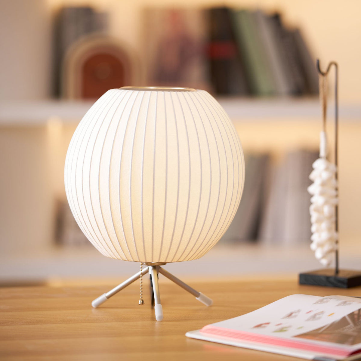 Soft focus bookshelves frame a round white Clérachic table lamp on a pale wooden desk, a pull chain hanging from its tripod base.