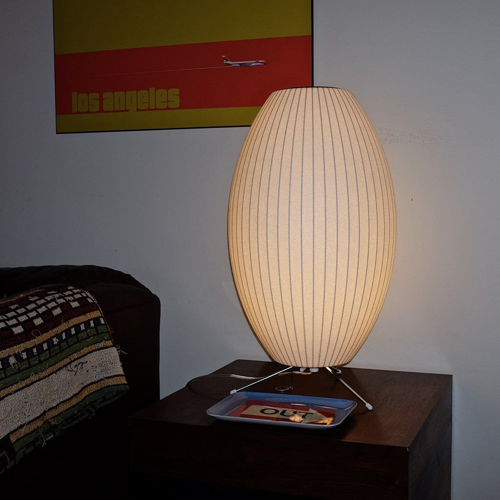 A tall, oval white Clérachic table lamp on a dark wooden side table, with a small tray at its feet and a poster behind.