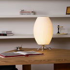An oval white Clérachic table lamp stands on a wooden table before white shelves, with an open book and a coffee cup beside it.