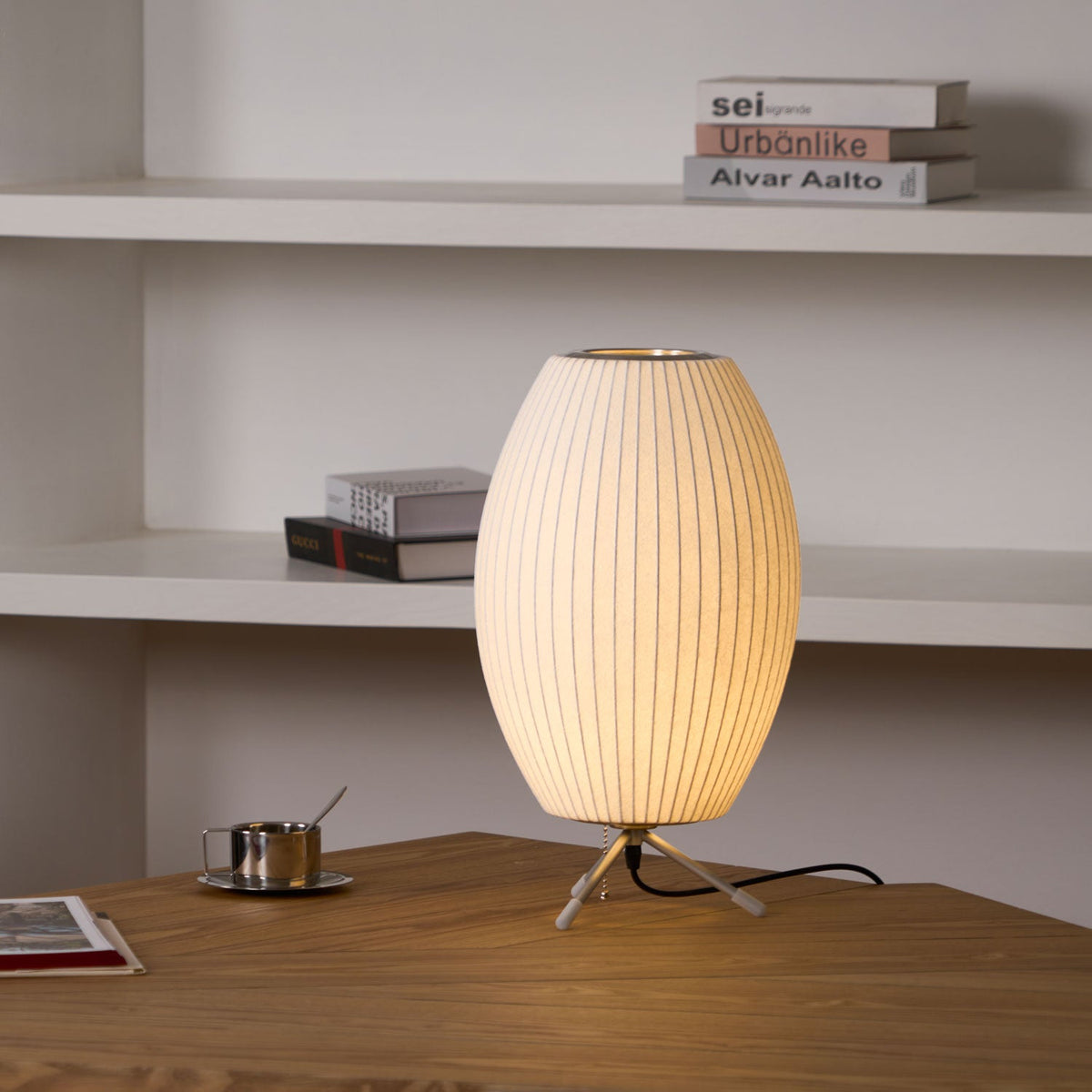 Closer on an oval white Clérachic table lamp, its tripod legs on a wooden table, with white shelves and stacked books behind.