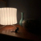 Close view of a tall, slightly waisted white shade on this Clérachic table lamp, glowing in a dim room beside a green vase.