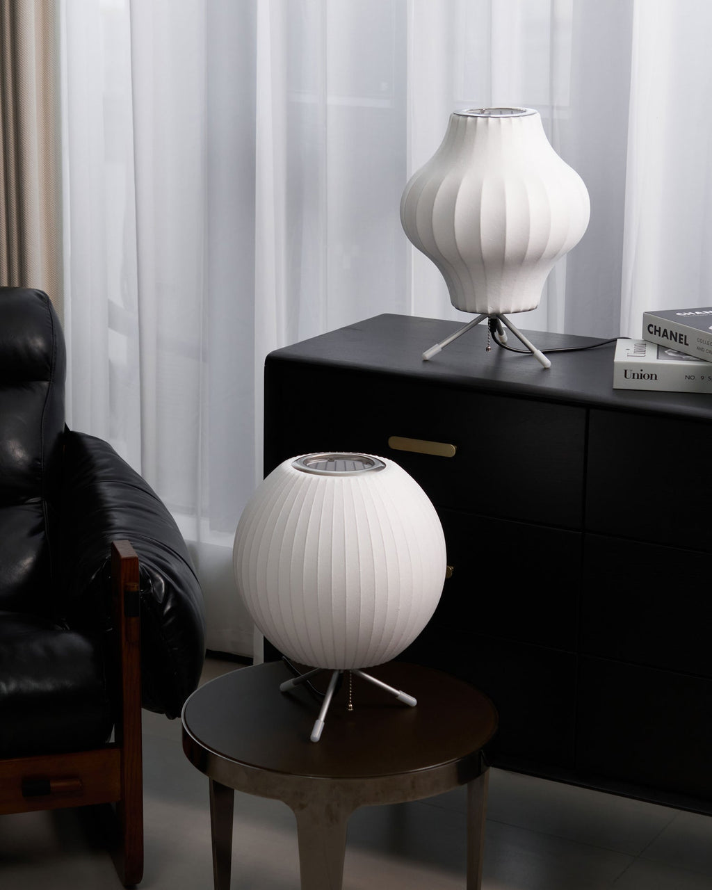 Two unlit white Clérachic table lamps, a pear shape on a black cabinet and a round one on a side table, by a black leather chair.