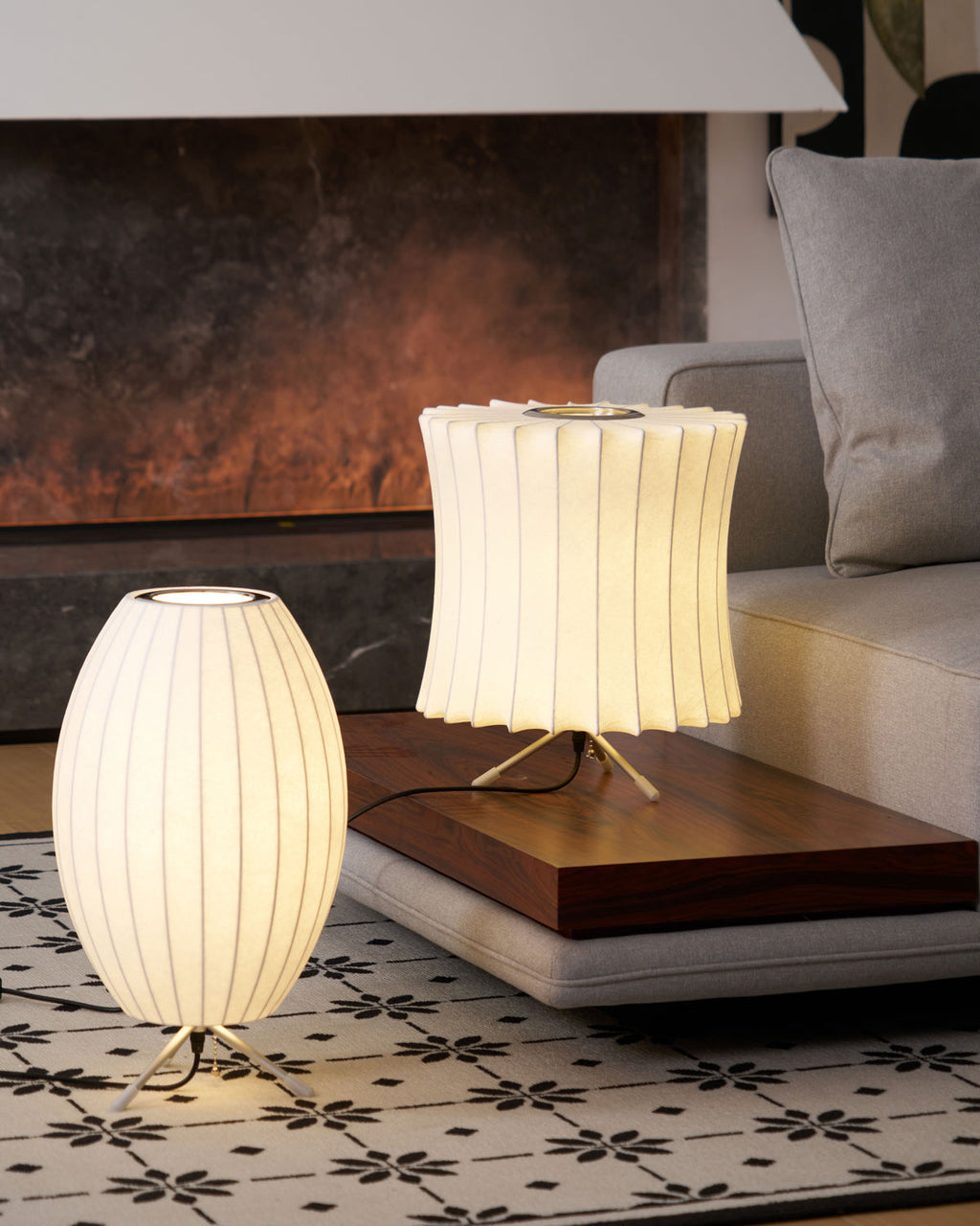 An oval and a waisted white Clérachic table lamp glow on a low wooden bench in front of a dark fireplace and a grey sofa.