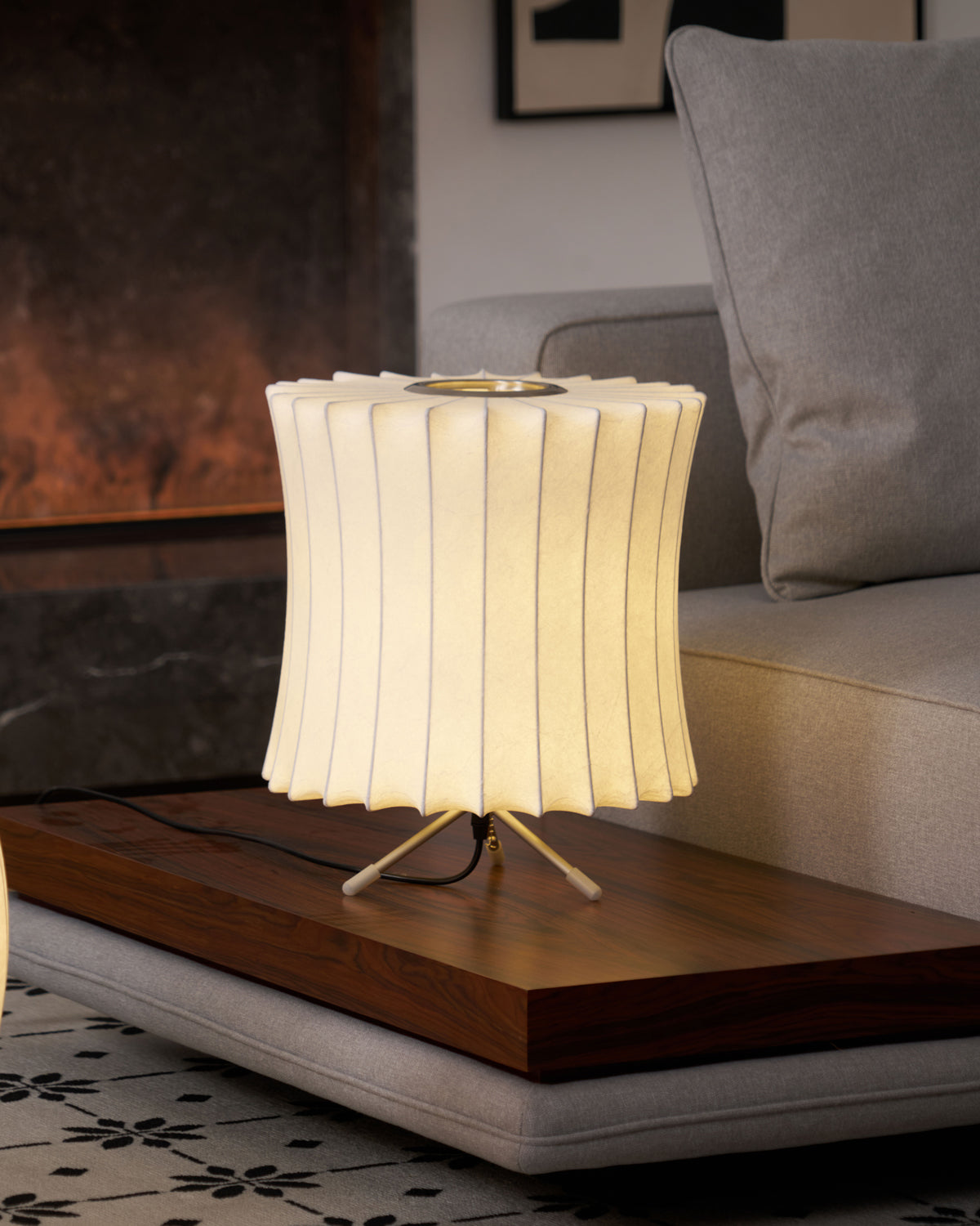 A waisted white Clérachic table lamp with a pleated top edge, lit on a low wooden bench beside a grey cushion and a sofa.
