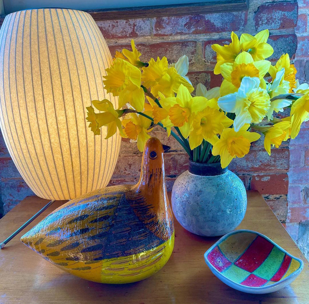 Close up of a large lit white Clérachic shade at the edge of the picture, with yellow daffodils, a blue-grey vase and a glass duck on a table.