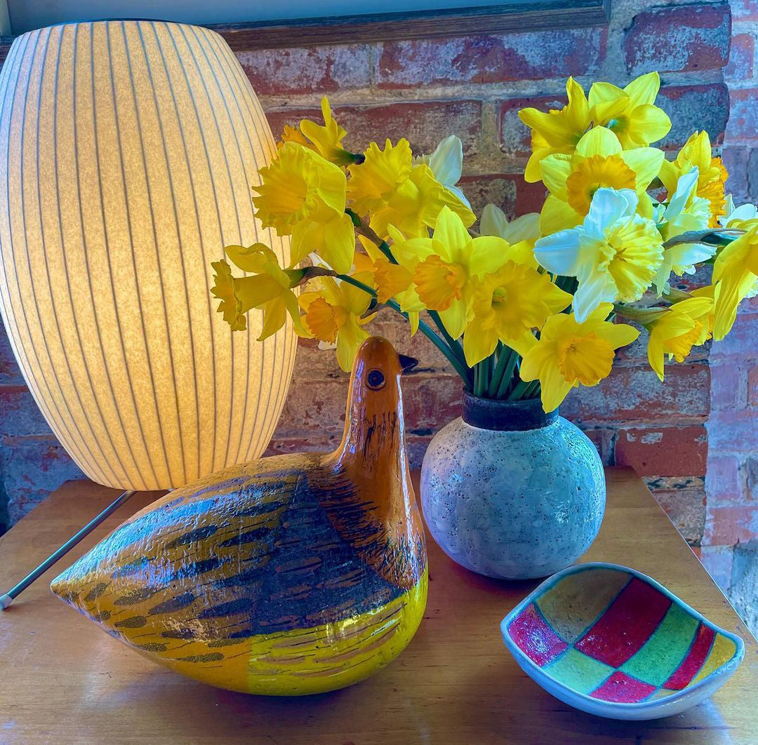 Close up of a large lit white Clérachic shade at the edge of the picture, with yellow daffodils, a blue-grey vase and a glass duck on a table.