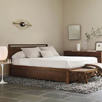 A white lamp glows on a small white side table beside a wooden bed in a calm bedroom, in this Clérachic room scene.