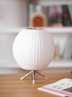 Round white ribbed Clérachic table lamp, unlit, on a pale wooden desk with an open magazine, against softly blurred shelves.