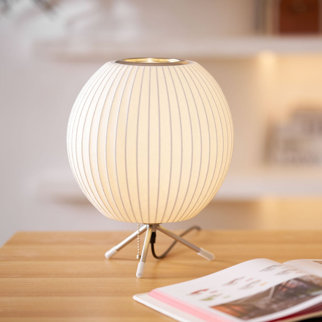 A round Clérachic table lamp, lit, on a light wooden desk beside an open magazine, with a pull chain by its tripod base.