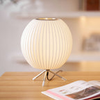 A round Clérachic table lamp, lit, on a light wooden desk beside an open magazine, with a pull chain by its tripod base.