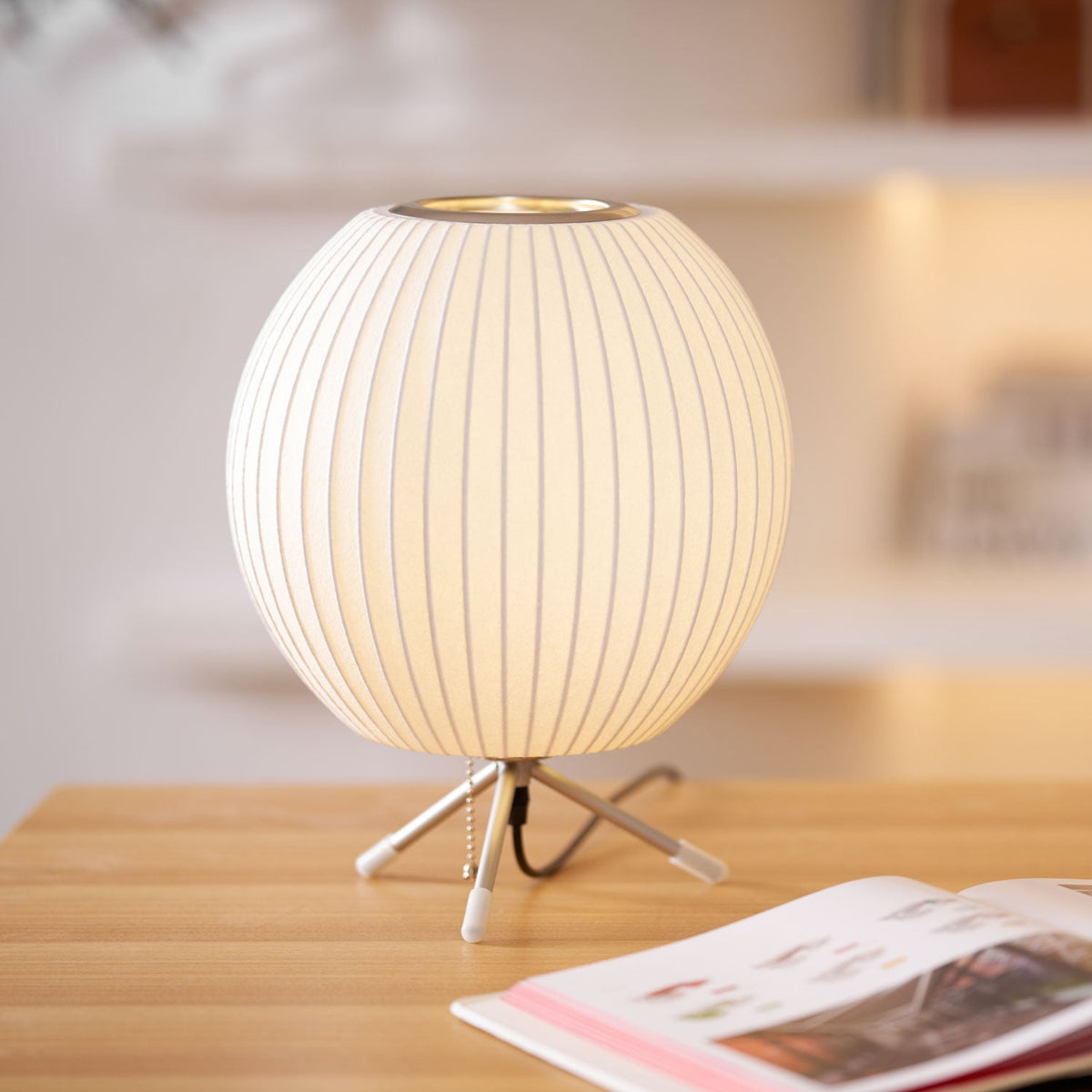 A round Clérachic table lamp, lit, on a light wooden desk beside an open magazine, with a pull chain by its tripod base.