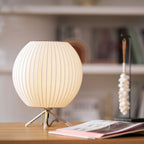 Glowing round Clérachic table lamp on a wooden desk, with a magazine in front and a white beaded ornament behind.