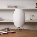 An unlit oval Clérachic table lamp on a wooden desk, with a coffee cup, an open book and white shelves behind it.