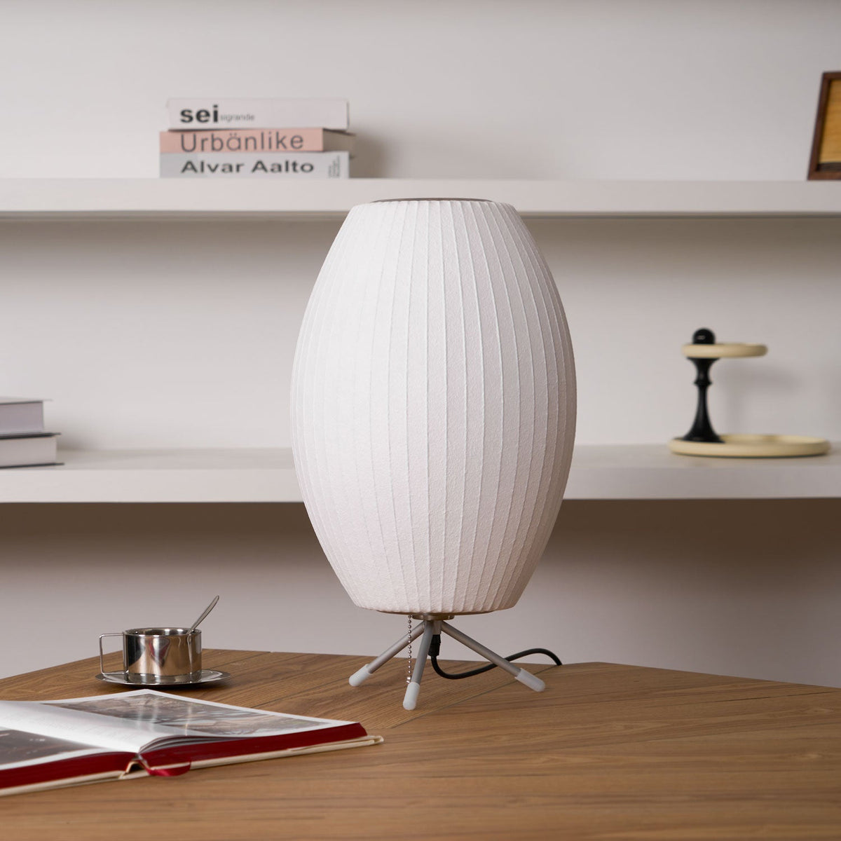 An unlit oval Clérachic table lamp on a wooden desk, with a coffee cup, an open book and white shelves behind it.
