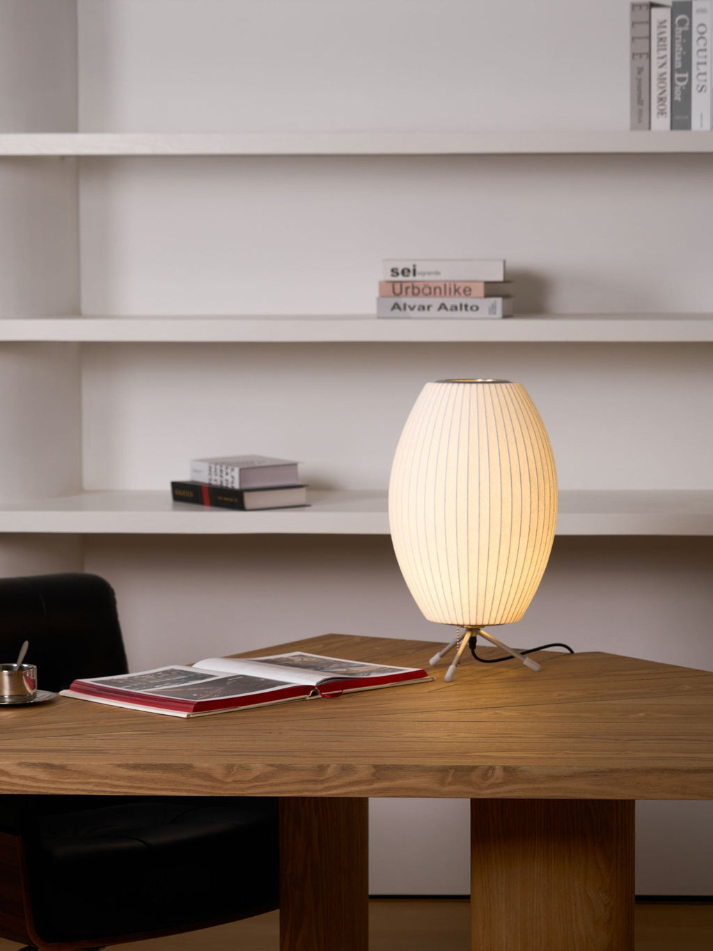 A lit oval white Clérachic table lamp on a wooden desk before open white shelves, with an open book beside it.