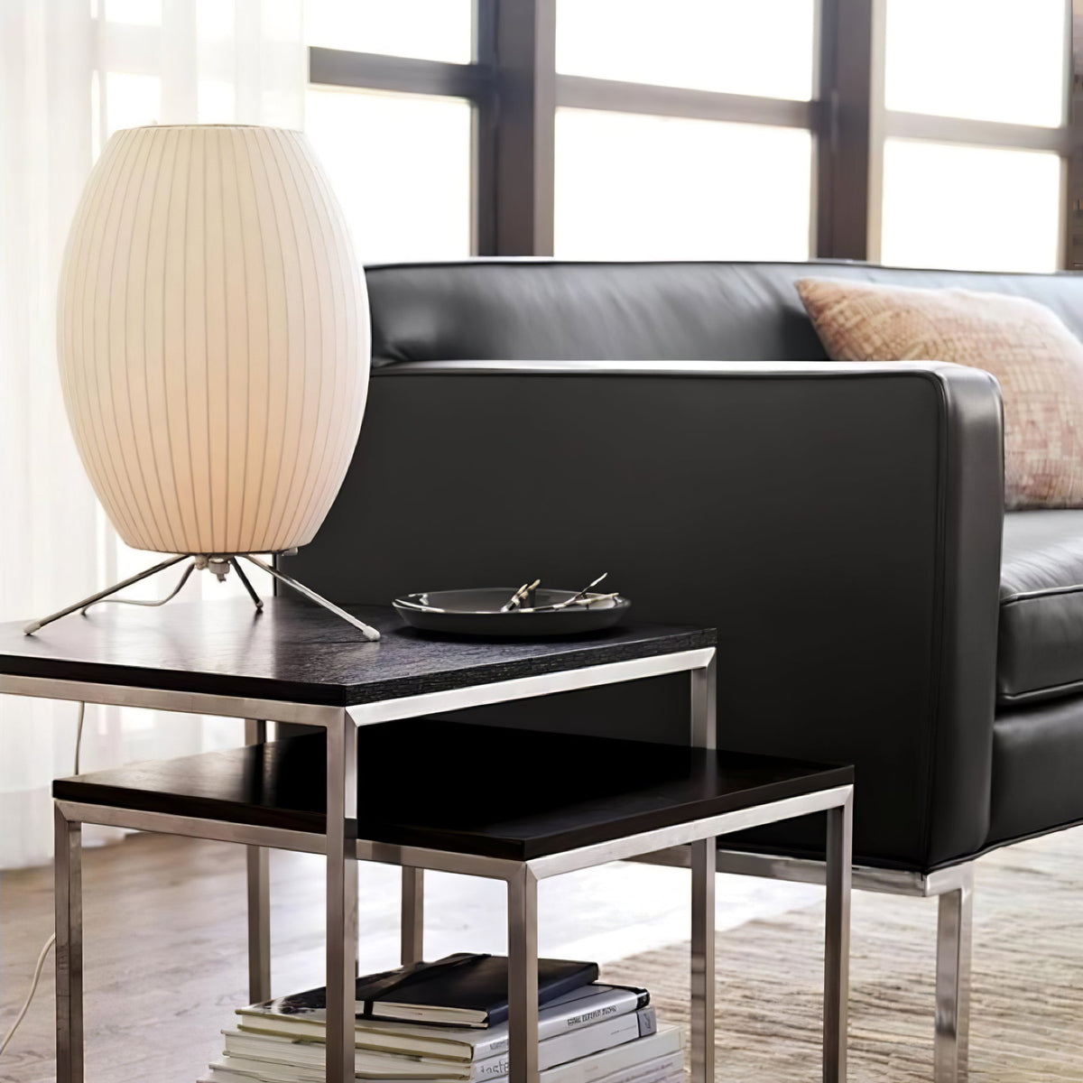A lit oval Clérachic table lamp on a black nesting side table beside a black leather sofa and large windows.
