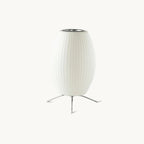 A white oval ribbed shade on a three-legged metal stand, this Clérachic table lamp seen on a plain pale background.