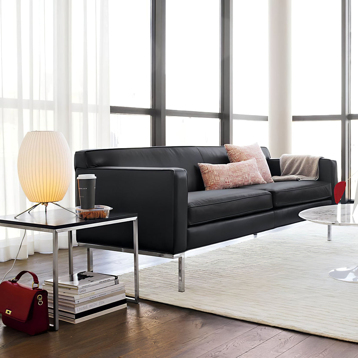 A small glowing oval Clérachic table lamp on a steel-framed side table beside a black leather sofa and floor-to-ceiling windows.