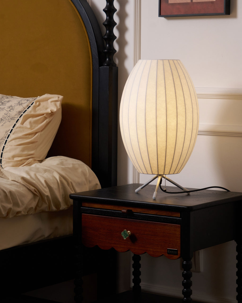 Oval Clérachic table lamp, lit, on a dark bedside table with a drawer, beside a bed with cream bedding and a mustard headboard.