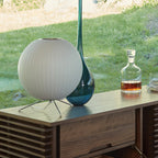 A round white Clérachic table lamp on a wooden sideboard, with a tall blue-green glass vase and a whisky decanter by a window.