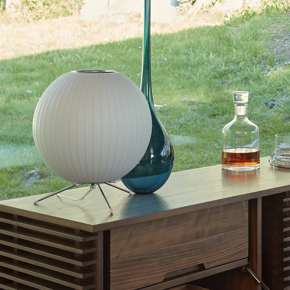 A round white Clérachic table lamp on a wooden sideboard, with a tall blue-green glass vase and a whisky decanter by a window.