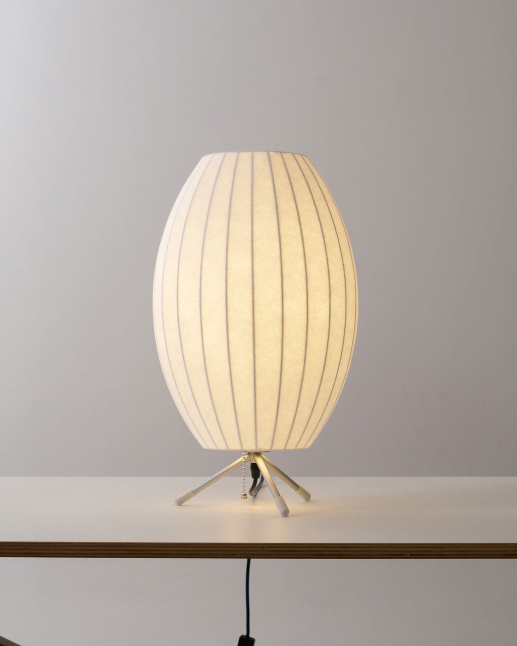 Oval white Clérachic table lamp lit on a white tabletop, its tripod legs catching the light against a grey wall.