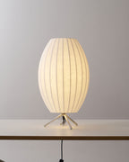 Oval white Clérachic table lamp lit on a white tabletop, its tripod legs catching the light against a grey wall.