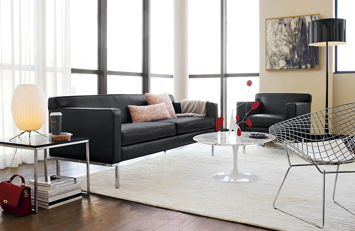 A small lit Clérachic table lamp on a side table beside a black leather sofa, with a white coffee table and a wire chair.