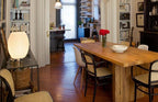 A lit Clérachic table lamp on a side table at the end of a long wooden dining table, in a bright room with bookshelves.