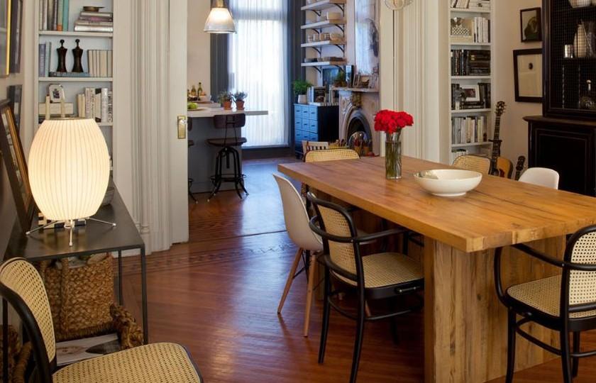 A lit Clérachic table lamp on a side table at the end of a long wooden dining table, in a bright room with bookshelves.