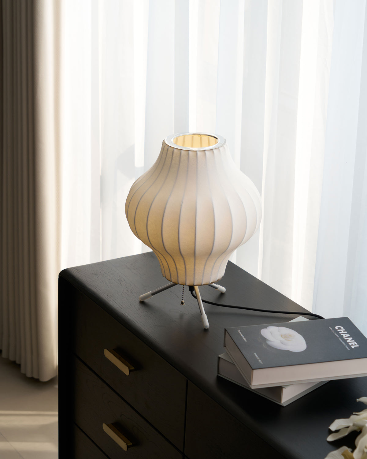 A pear-shaped Clérachic table lamp, lit, on a black chest with brass handles, beside an art book and a few white flowers.