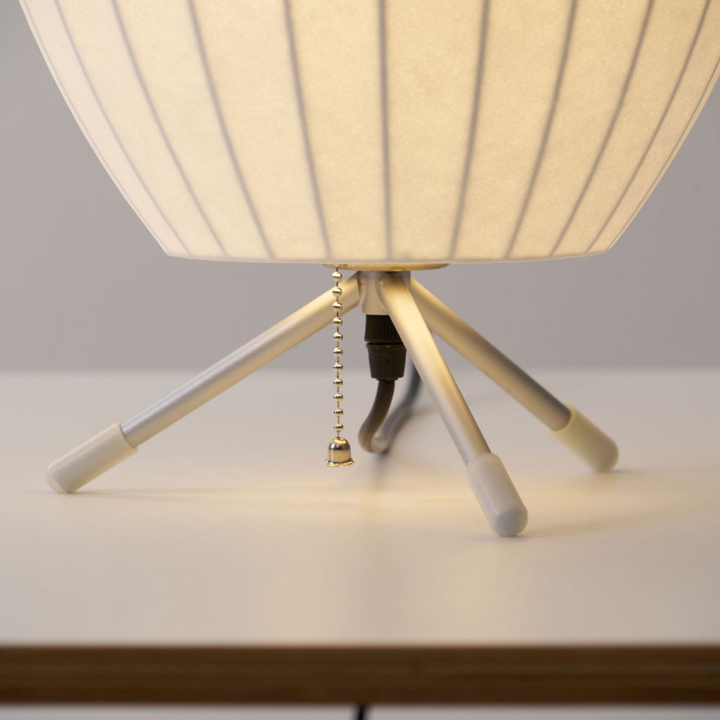 The slim tripod legs, white tips, pull chain and black cord beneath the round shade of this Clérachic table lamp.