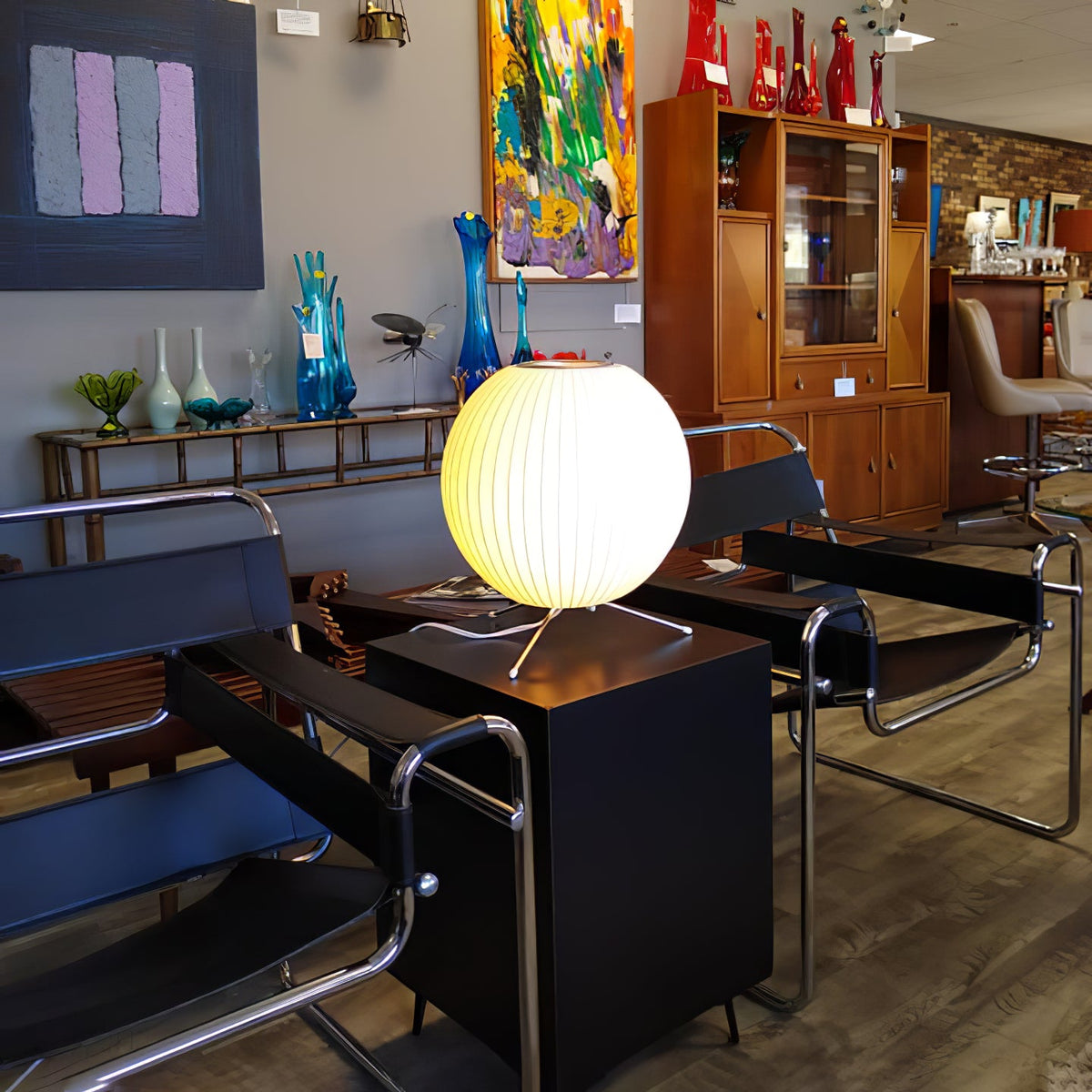 A round, lit Clérachic table lamp on a black cabinet in a shop with chrome chairs, blue glass vases and colourful art.