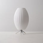 Unlit oval white Clérachic table lamp on a grey tabletop and backdrop, its slim tripod legs and black cord visible.