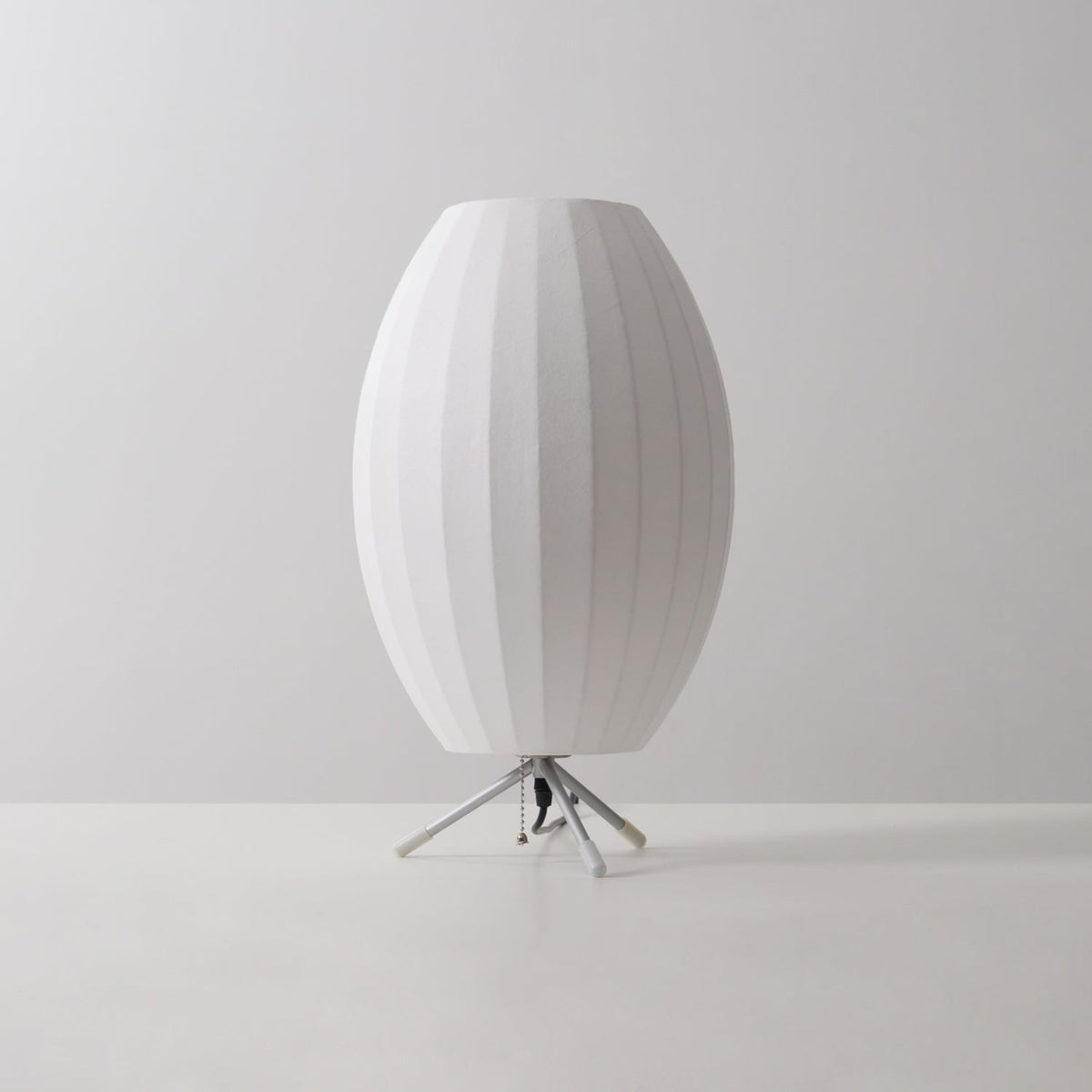 Unlit oval white Clérachic table lamp on a grey tabletop and backdrop, its slim tripod legs and black cord visible.