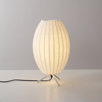 A lit oval Clérachic table lamp on a grey backdrop, a warm glow at its tripod base and a black cord trailing to the left.