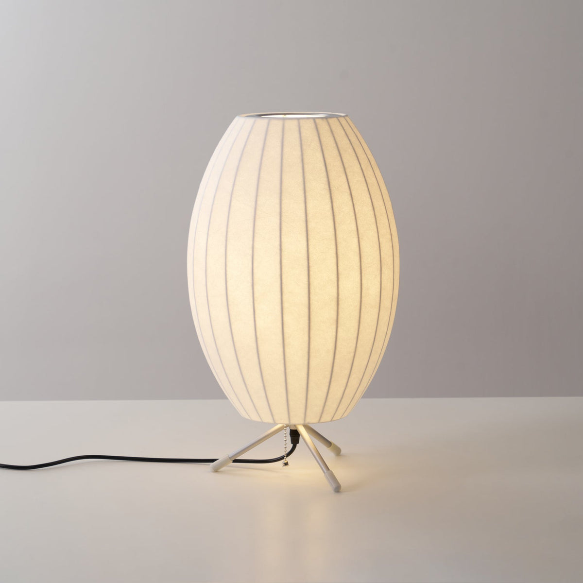 A lit oval Clérachic table lamp on a grey backdrop, a warm glow at its tripod base and a black cord trailing to the left.