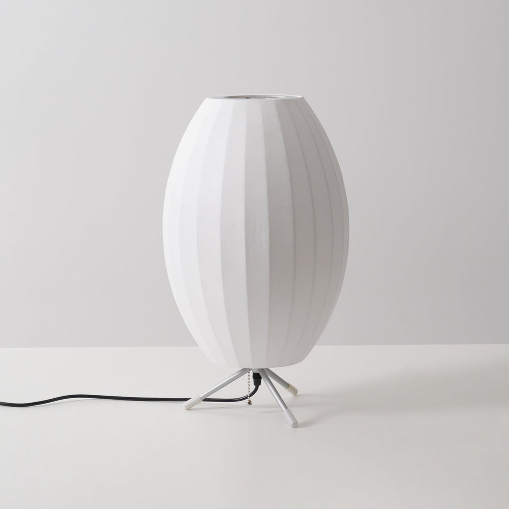 An unlit oval white shade with a metal ring at the top, this Clérachic table lamp is shown at a slight angle on a grey set.