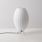 An unlit oval white shade with a metal ring at the top, this Clérachic table lamp is shown at a slight angle on a grey set.