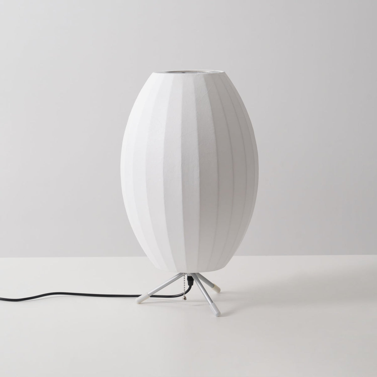 An unlit oval white shade with a metal ring at the top, this Clérachic table lamp is shown at a slight angle on a grey set.